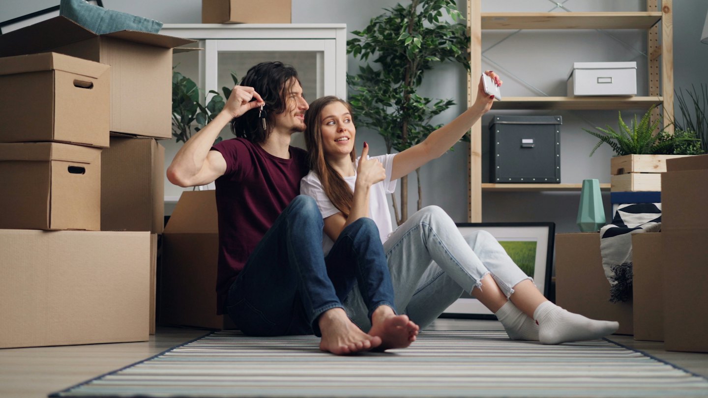 Couple takes a selfie with keys in their new home surrounded by boxes.