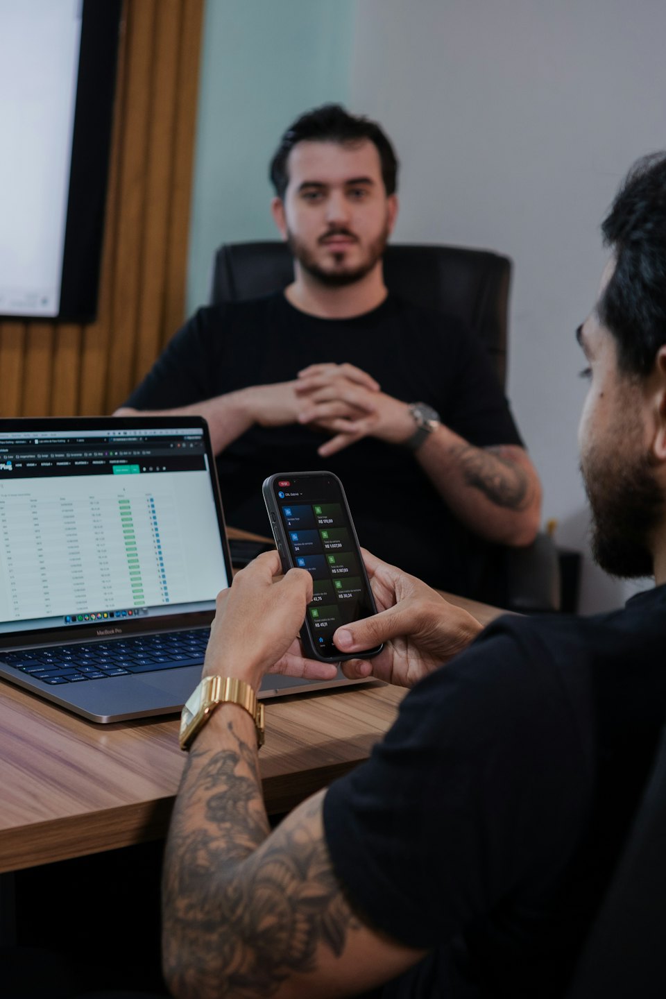 Two men discuss business data at a wooden office desk.